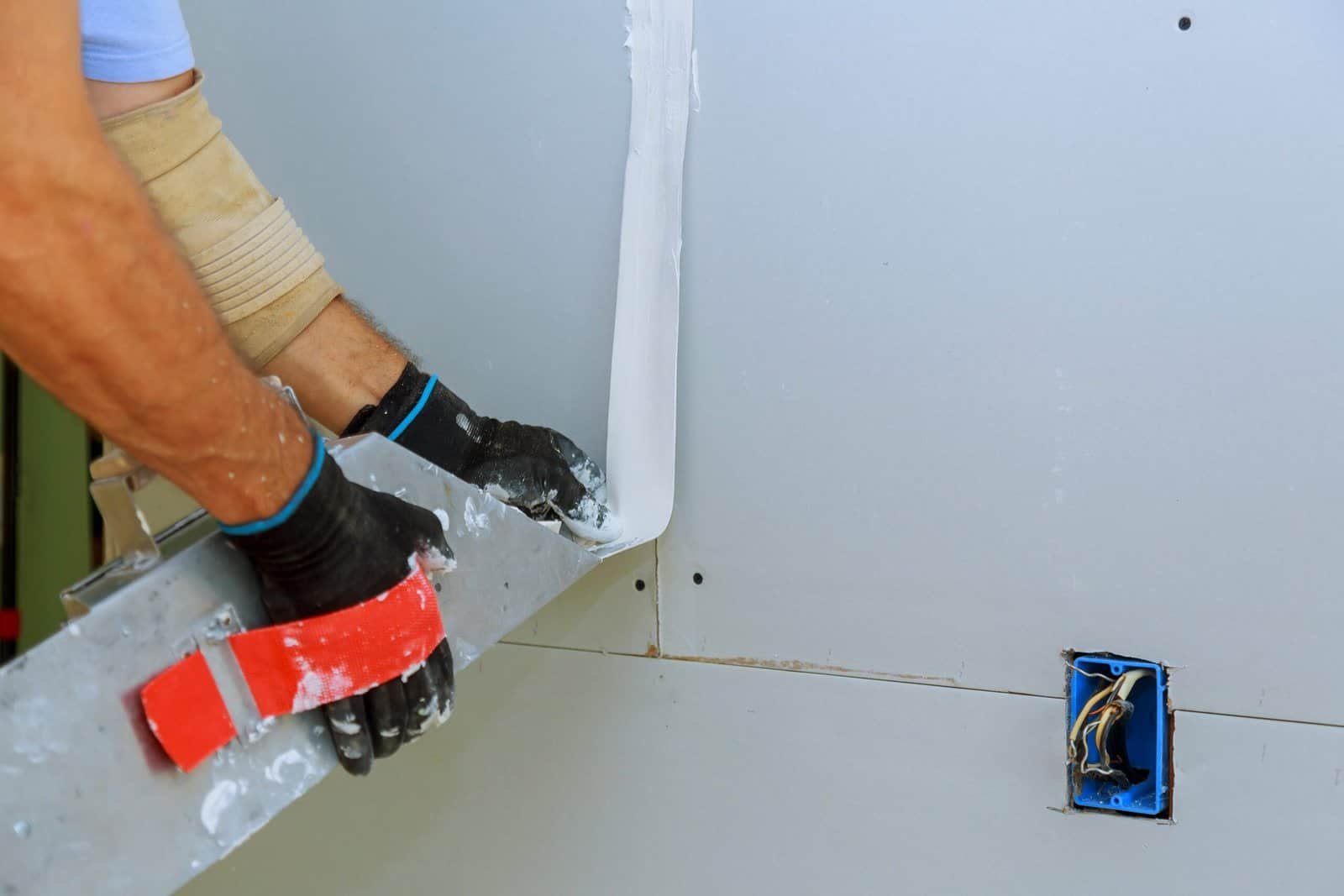 Worker installing drywall with joint compound and wiring in an electrical box.
